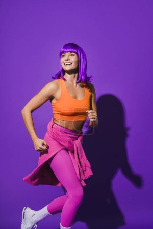 Foto de Joven mujer alegre con ropa deportiva de colores corriendo sobre fondo púrpura - Imagen libre de derechos