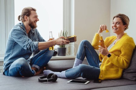 Téléchargez les photos : Jeune couple joyeux assis sur le canapé boire de la bière et manger des nachos à la maison - en image libre de droit