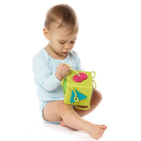 Téléchargez les photos : Adorable petit garçon dans une barboteuse est assis et jouer avec un jouet en plastique sur fond blanc. - en image libre de droit