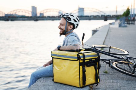 Photo for Young smiling express delivery courier is sitting on a quay and looking at the quiet river water with insulated bag and bicycle nearby. - Royalty Free Image