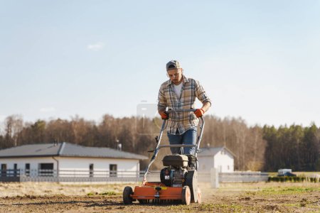 Téléchargez les photos : Jeune homme utilisant une machine d'aération pour scarifier et aérer la pelouse ou le pré - en image libre de droit