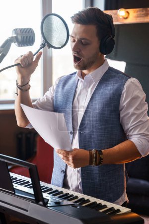 Téléchargez les photos : Jeune homme barbu beau chante dans un microphone dans un studio de musique maison en face d'un piano avec un miroir de maquillage derrière son dos. - en image libre de droit