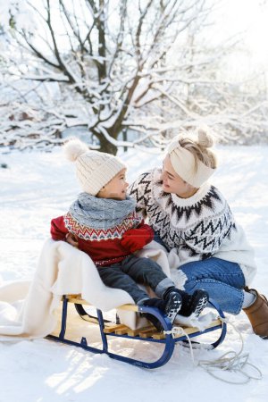 Téléchargez les photos : Jeune mère et son mignon petit fils avec traîneau rétro dans un parc enneigé pendant la journée ensoleillée d'hiver - en image libre de droit