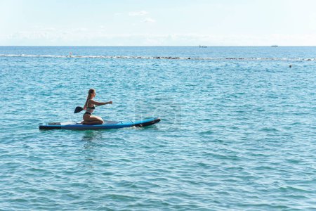 Foto de Joven surfista femenina sexy en bikini está montando un stand-up paddleboard y remando con una paleta en un océano. - Imagen libre de derechos
