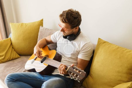 Foto de Joven barbudo con gafas sentado en el sofá está tocando casualmente la guitarra acústica en una habitación iluminada. - Imagen libre de derechos
