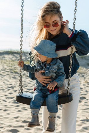 Photo for Young smiling mother is holding her little son sitting on a swing at the sandy beach. - Royalty Free Image