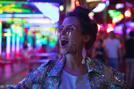 Téléchargez les photos : Jeune femme élégante portant une veste avec des paillettes brillantes sur la rue de la ville avec des néons - en image libre de droit