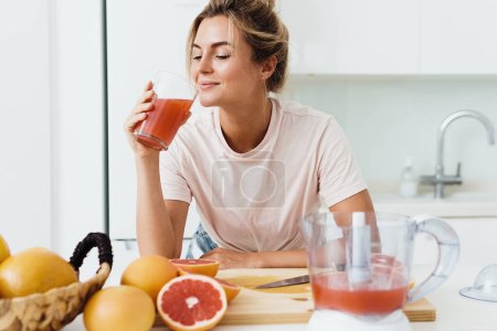 Foto de Mujer joven bebiendo zumo de pomelo casero recién exprimido en cocina blanca - Imagen libre de derechos