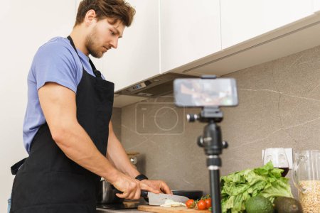 Photo for Young bearded chef blogger is cutting onions with a knife on a cutting board in a kitchen in front of the camera. - Royalty Free Image