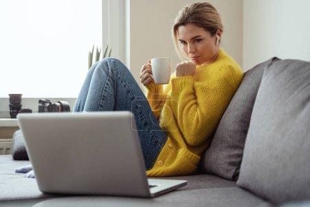 Jeune femme avec tasse de café chaud assis sur le canapé et à l'aide d'un ordinateur portable à la maison