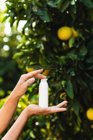 Foto de Mujer sostiene una botella blanca de producto para el cuidado de la piel en sus manos sobre fondo borroso de limonero. - Imagen libre de derechos