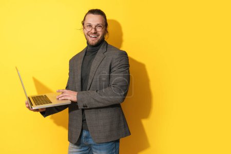 Foto de Guapo hombre barbudo feliz con anteojos está utilizando ordenador portátil sobre fondo amarillo - Imagen libre de derechos