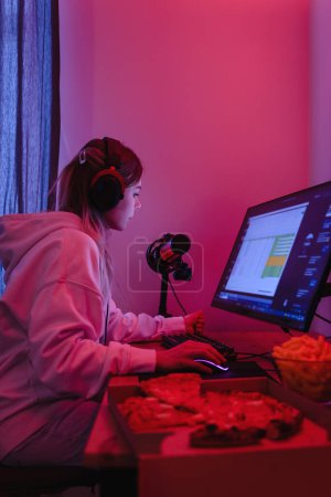 Foto de Mujer joven gamer o desarrollador de software sentado en la computadora personal moderna y comer comida chatarra por la noche en la habitación con luces de neón - Imagen libre de derechos