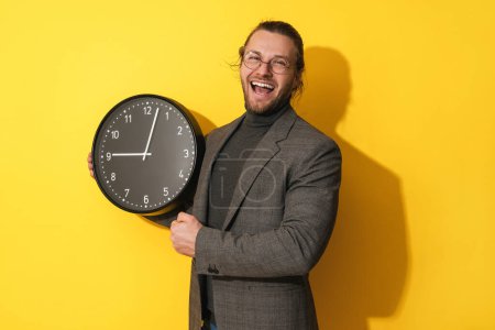 Foto de Hombre barbudo alegre con gafas sosteniendo gran reloj sobre fondo amarillo - Imagen libre de derechos