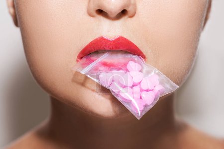 Foto de Primer plano de una mujer joven sosteniendo una bolsa de cierre transparente llena de píldoras en forma de corazón rosa en su boca. - Imagen libre de derechos