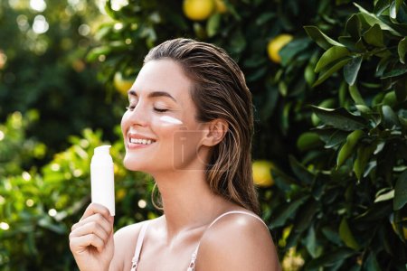 Téléchargez les photos : Portrait extérieur d'une femme joyeuse et belle qui applique de la crème hydratante ou de la crème solaire sur sa peau faciale - en image libre de droit