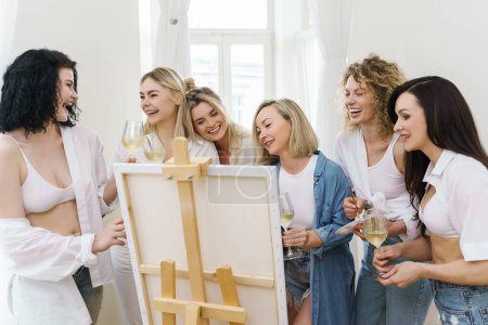 Foto de Grupo de mujeres jóvenes hermosas pintan sobre lienzo y beben vino blanco durante la fiesta en casa - Imagen libre de derechos