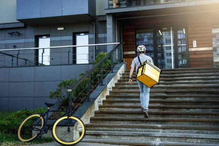 Téléchargez les photos : Jeune messager express livraison de nourriture avec sac isolé derrière son dos monte les escaliers après le stationnement de son vélo. - en image libre de droit