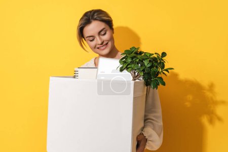 Téléchargez les photos : Jeune femme heureuse tenant boîte avec des objets personnels après la promotion de l'emploi sur fond jaune - en image libre de droit