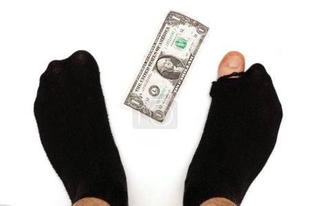 Photo for Closeup shot of a male feet in a black hoaly socks with sticking out toe and a one dollar banknote on white background. Concept of poverty and financial crisis. - Royalty Free Image