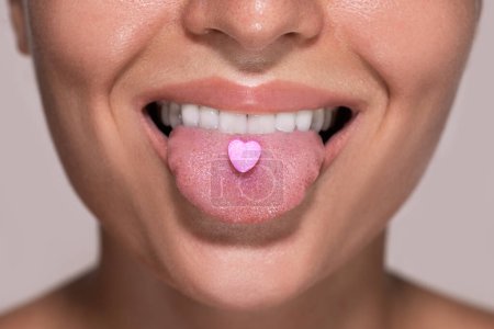 Foto de Primer plano de una parte inferior de una cara femenina joven con una píldora rosa en forma de corazón en su lengua. - Imagen libre de derechos