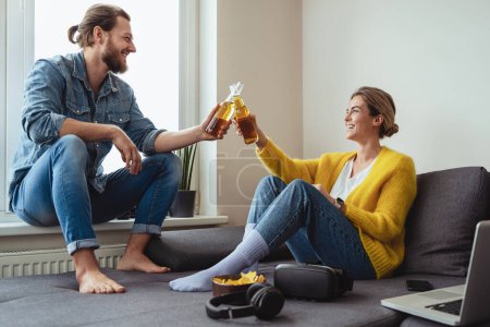 Foto de Joven pareja alegre sentada en el sofá bebiendo cerveza y comiendo nachos en casa - Imagen libre de derechos