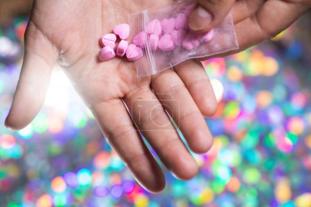Foto de Primer plano de un hombre vertiendo píldoras en forma de corazón rosa en su palma de una bolsa de cierre transparente sobre un fondo borroso brillante. - Imagen libre de derechos