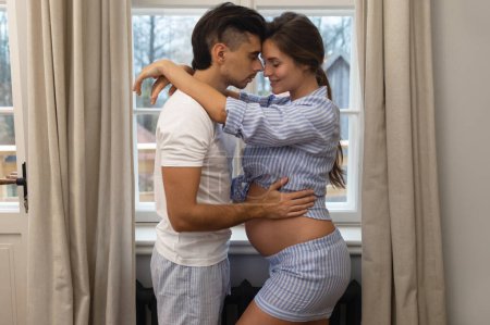 Foto de Una joven pareja sensual está esperando un bebé. Hombre y su esposa embarazada en abrazo al lado de la ventana. - Imagen libre de derechos