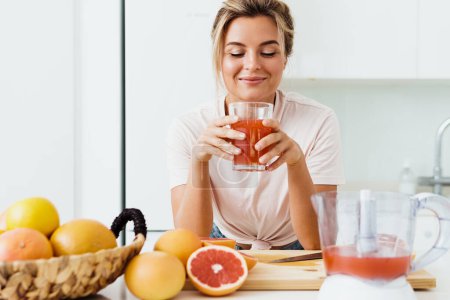 Photo for Young woman drinking freshly squeezed homemade grapefruit juice in white kitchen - Royalty Free Image