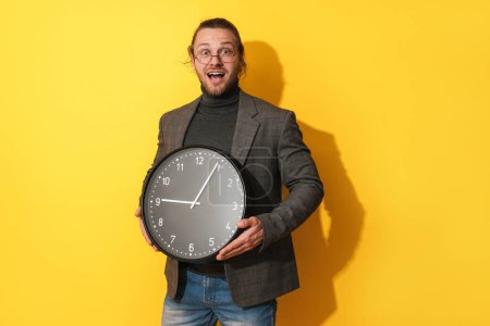 Photo for Surprised bearded man wearing glasses holding big clock on yellow background - Royalty Free Image