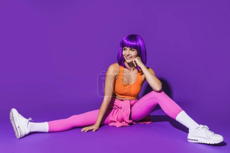 Téléchargez les photos : Portrait de femme joyeuse portant des vêtements de sport colorés assis sur fond violet - en image libre de droit