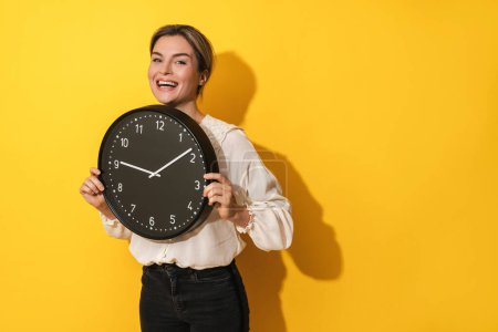Photo for Cheerful young woman holding big clock on yellow background - Royalty Free Image
