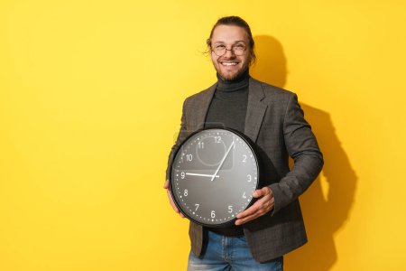Photo for Cheerful bearded man wearing glasses holding big clock on yellow background - Royalty Free Image