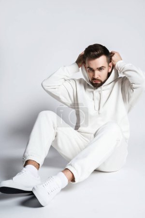 Foto de Hombre guapo con capucha blanca en blanco y pantalones sentados sobre fondo gris claro - Imagen libre de derechos