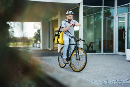 Photo for Young express food delivery courier riding bicycle with insulated bag behind his back is looking at his watch. - Royalty Free Image