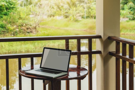 Foto de Ordenador portátil con pantalla en blanco de pie sobre una pequeña mesa de madera en una terraza de una cabaña en una zona rural tranquila. - Imagen libre de derechos