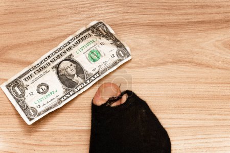 Foto de Primer plano de un pie masculino en un calcetín negro con puntera sobresaliente y un billete de un dólar sobre fondo de madera. Concepto de pobreza y crisis financiera. - Imagen libre de derechos