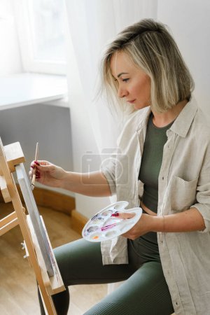 Téléchargez les photos : Jeune femme artiste peinture sur toile sur le chevalet à la maison studio - en image libre de droit
