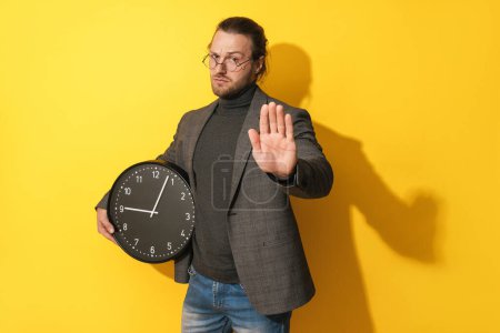 Photo for Serious man wearing glasses holding clock and showing stop gesture on yellow background - Royalty Free Image