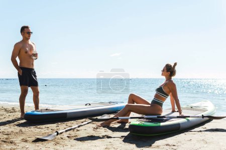 Téléchargez les photos : Les jeunes surfeurs de paddleboard adultes mâles et femelles se détendent et bronzent sur une plage de sable près de l'océan. - en image libre de droit