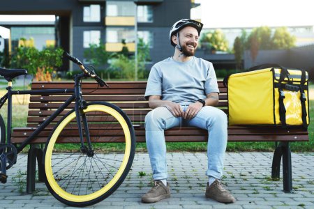Photo for Young smiling express delivery courier is sitting and resting on a bench with insulated bag and bicycle nearby. - Royalty Free Image