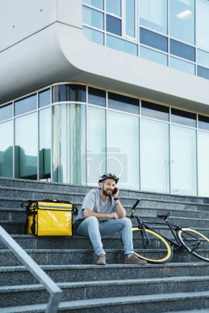 Foto de Joven sonriente mensajero de reparto de comida express está sentado en las escaleras con bolsa aislante y bicicleta y hablando por su teléfono. - Imagen libre de derechos