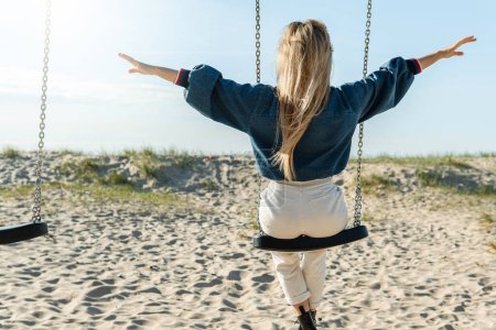 Téléchargez les photos : Plan arrière d'une jeune femme se balançant sur la balançoire et étendant les mains sur la plage de sable. - en image libre de droit