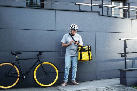 Foto de Joven mensajero de reparto de comida express con bolsa aislada en su lado está mirando su teléfono cerca de la bicicleta estacionada. - Imagen libre de derechos