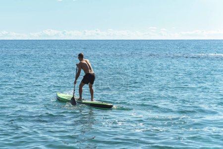 Photo for Young male surfer is riding a standup paddleboard and rowing with a paddle in an ocean. - Royalty Free Image