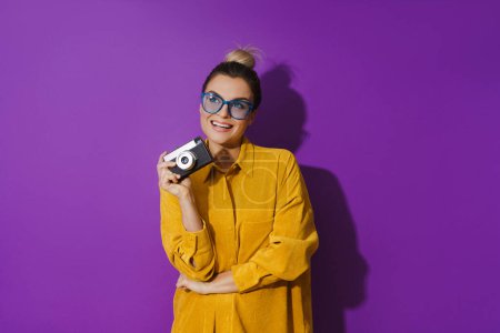 Photo for Portrait of young cheerful girl wearing eyeglasses holding vintage film camera on purple background - Royalty Free Image