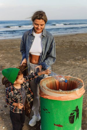 Téléchargez les photos : La famille caucasienne collecte des déchets plastiques sur la plage. Mère enseigne à son fils à garder la nature propre. - en image libre de droit