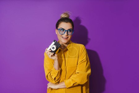 Téléchargez les photos : Portrait de jeune fille joyeuse portant des lunettes tenant caméra de film vintage sur fond violet - en image libre de droit