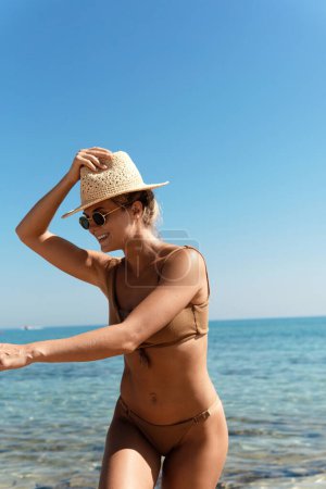 Téléchargez les photos : Belle femme insouciante portant un bikini brun et un chapeau de paille pendant ses vacances à la plage près de la mer bleue. - en image libre de droit