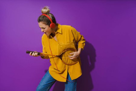 Téléchargez les photos : Fille joyeuse se bloque dans sa chemise jaune tout en portant des écouteurs rouges. Elle se balade sur sa raquette comme si c'était une guitare, sur un fond violet vibrant. - en image libre de droit